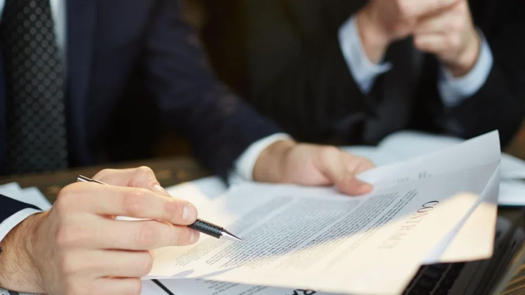 Mão segurando caneta sobre um documento de contrato, com outra mão segurando uma folha, representando a assinatura de acordos de recuperação judicial