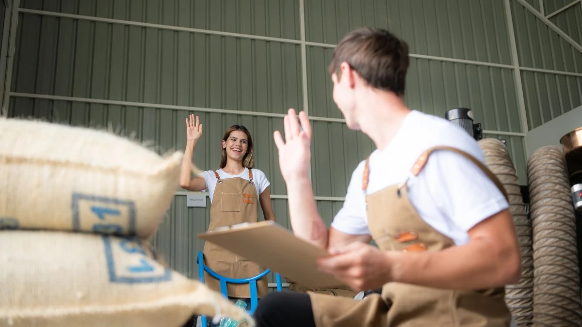 Imagem de jovens trabalhadores em revenda agrícola, sorrindo e acenando um para o outro enquanto seguram prancheta e estão cercados por sacos de material.