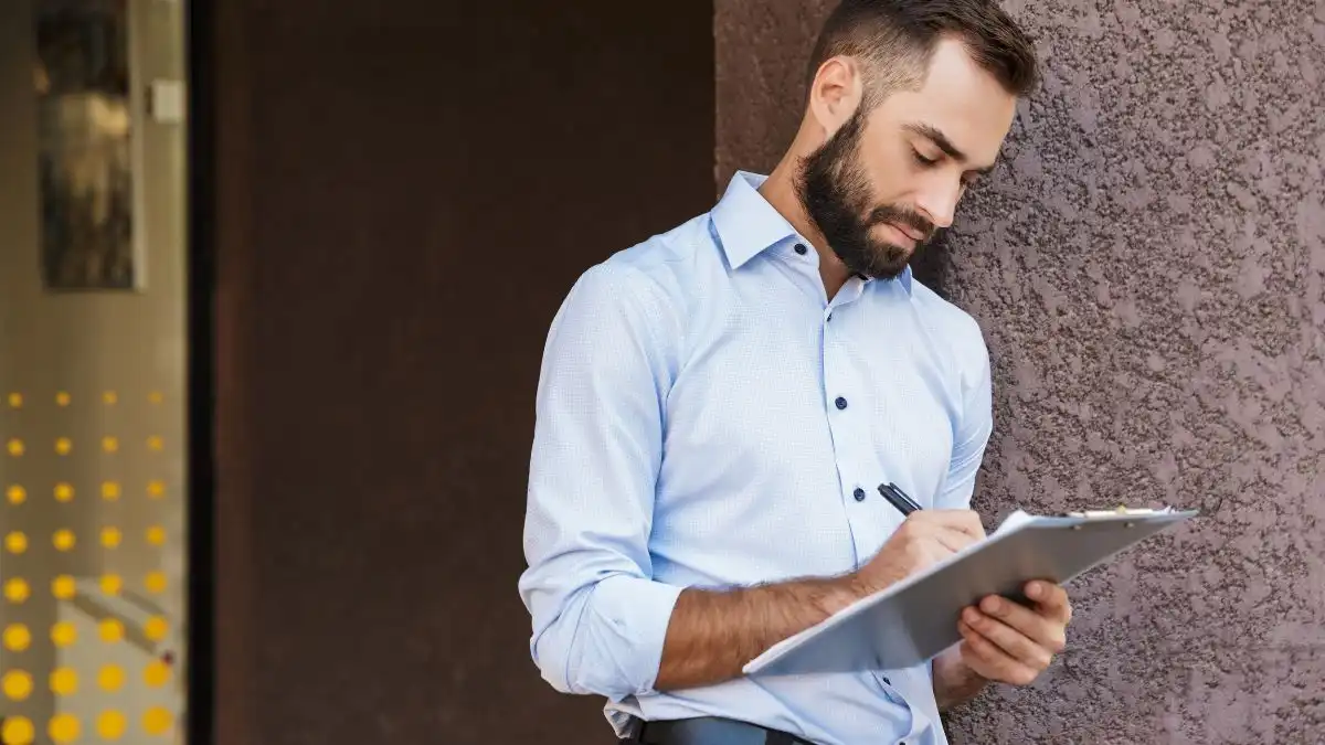 Homem de camisa azul escrevendo em um bloco de notas, com expressão concentrada e apoiado em uma parede. Ideal para ilustrar atividades de planejamento ou trabalho.